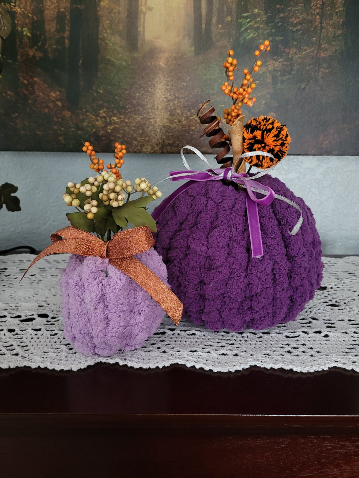 Two decorative knitted pumpkins in purple and lavender tones with faux floral arrangements and autumn accents, displayed on a white lace doily in front of a forest backdrop.